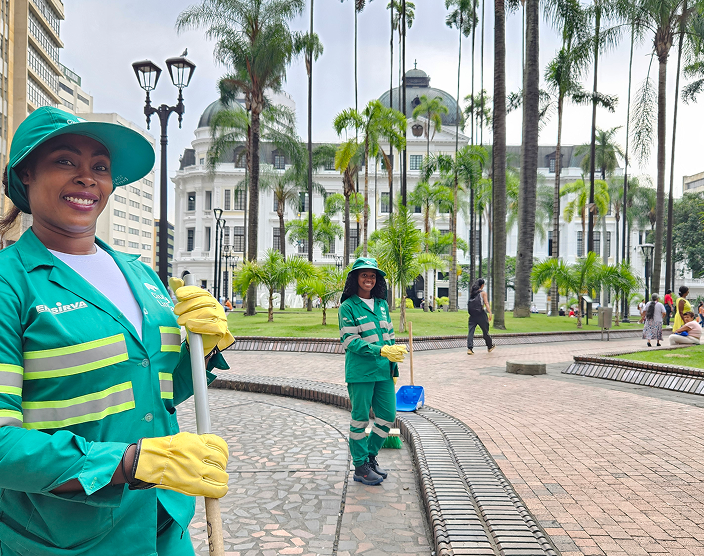 Dos colaboradoras de Ciudad Limpia realizan labores de limpieza en un espacio público con palmas y senderos peatonales, frente a una edificación emblemática del centro urbano.