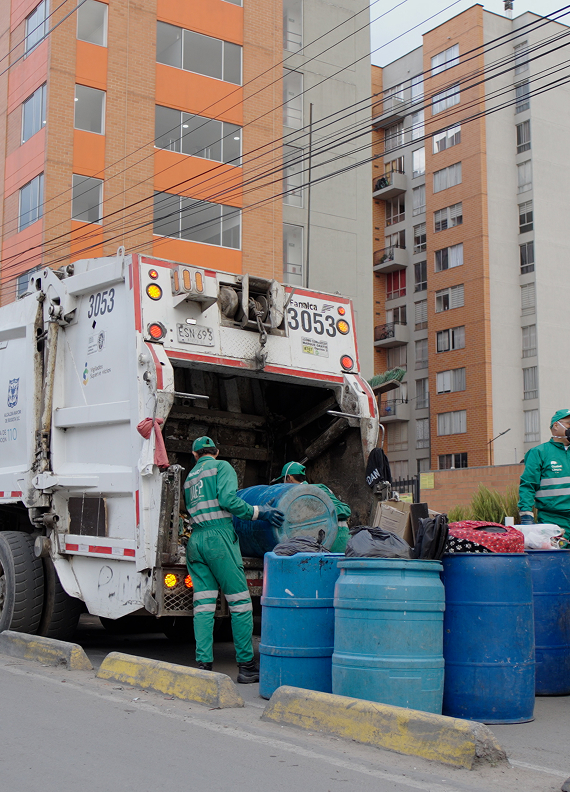Colaboradores de Ciudad Limpia realizando la recolección de residuos junto a un camión compactador, mientras cargan contenedores azules en una vía urbana rodeada de edificios residenciales.