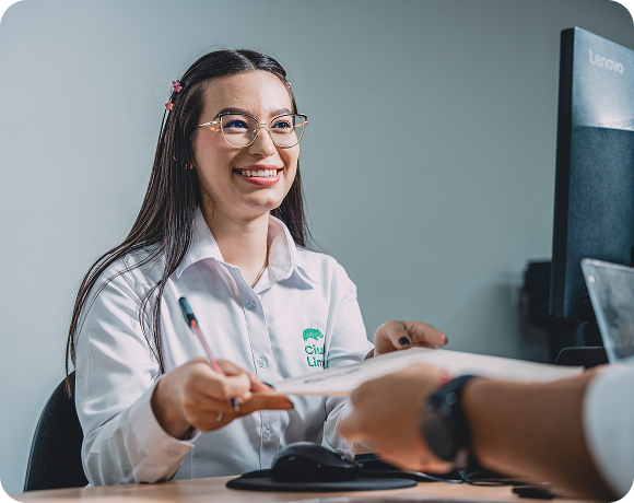 Colaboradora de Ciudad Limpia atiende a una persona en un punto de servicio y le entrega un documento, con una expresión amable frente al computador.