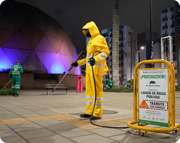 Colaborador de Ciudad Limpia realizando lavado de áreas públicas durante una jornada nocturna, con traje impermeable amarillo, equipo de aspersión y señalización preventiva en el espacio intervenido.