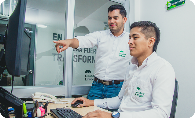 Dos colaboradores de Ciudad Limpia trabajan frente a un computador en una oficina, mientras uno señala la pantalla en una dinámica de acompañamiento y revisión conjunta.