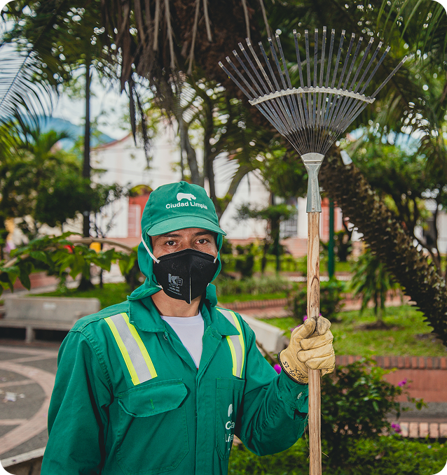 Fotografía de un colaborador de Ciudad Limpia con uniforme verde, guantes y mascarilla posando en una zona verde urbana, mientras sostiene un rastrillo de jardinería; al fondo se observan palmeras, senderos y áreas con jardines.