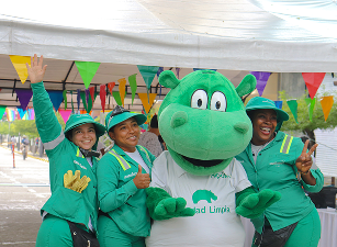 Tres colaboradoras de Ciudad Limpia posando sonrientes junto a Hipólito, la mascota institucional en una actividad comunitaria, bajo una carpa decorada con banderines de colores.