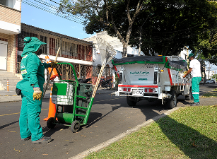 Colaboradora de Ciudad Limpia permanece junto a un carro de apoyo con herramientas de barrido, mientras su compañero trabaja cerca de un vehículo de servicio en una vía urbana.