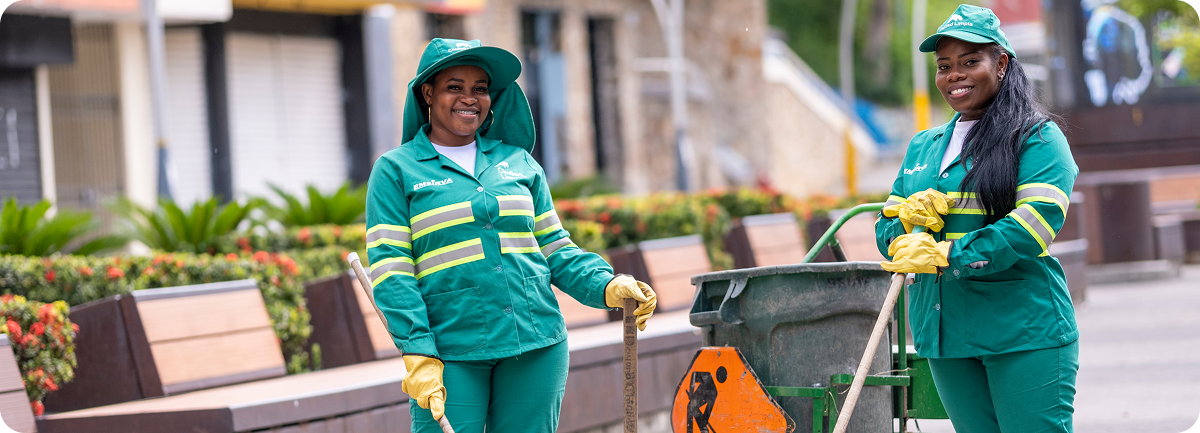 Dos colaboradoras de Ciudad Limpia, con uniforme verde reflectivo y guantes amarillos, posan sonrientes junto a un carro de aseo y herramientas de limpieza en un espacio público urbano. Al fondo se observan bancas, jardines ornamentales y una zona peatonal en mantenimiento.