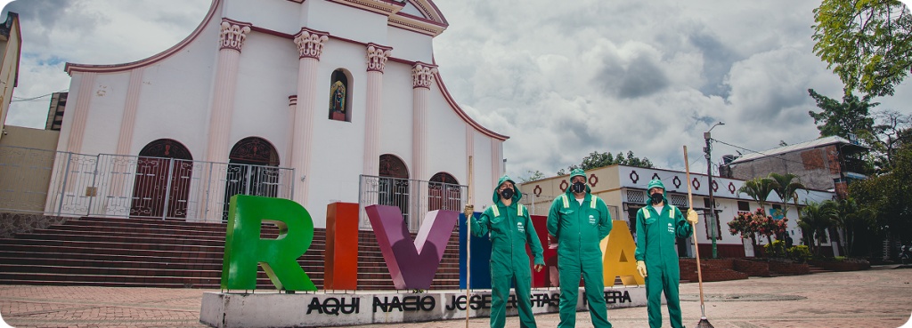 Tres colaboradores de Ciudad Limpia, con uniforme verde, guantes y mascarilla, posan con escobas frente al letrero turístico de “RIVERA” en una plaza principal. Al fondo se observa una iglesia de fachada blanca con detalles rosados, escaleras y edificaciones del entorno urbano bajo un cielo nublado.