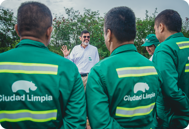 Grupo de colaboradores de Ciudad Limpia participando en una charla al aire libre. En primer plano se observan varios trabajadores de espaldas con uniformes verdes reflectivos, mientras al centro un directivo o coordinador, con camisa blanca institucional, dirige la conversación frente a una zona verde.