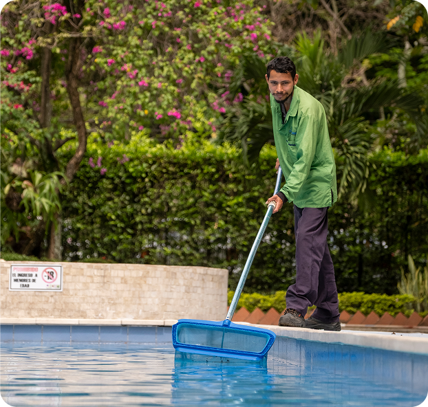 Colaborador de Ciudad Limpia realizando la limpieza de una piscina con una red de mango largo, retirando residuos de la superficie del agua. La escena transcurre en una zona residencial con jardines, arbustos y árboles con flores alrededor.