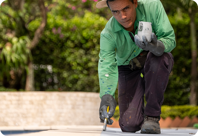Colaborador de Ciudad Limpia realizando labores de mantenimiento en el borde de una piscina, aplicando pintura o recubrimiento con una brocha mientras sostiene un recipiente con material. Viste uniforme verde, guantes y sombrero, y trabaja en un entorno exterior rodeado de zonas verdes.