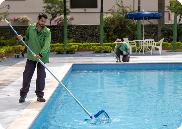 Dos colaboradores de Ciudad Limpia realizan labores de mantenimiento en la zona de piscina de un conjunto residencial. En primer plano, uno de ellos limpia el agua con una red de mango largo, mientras al fondo otro trabajador adelanta tareas de aseo en el borde de la piscina, rodeada de jardines y mobiliario exterior.