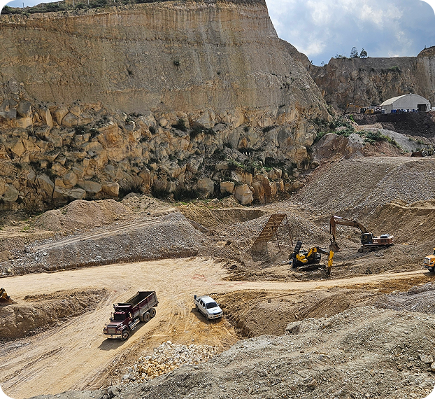 Vista panorámica de una cantera o zona rocosa a cielo abierto, con montículos de material y maquinaria amarilla en operación. En la parte baja se observan una volqueta, una camioneta y equipos de movimiento de tierra en plena jornada de trabajo.