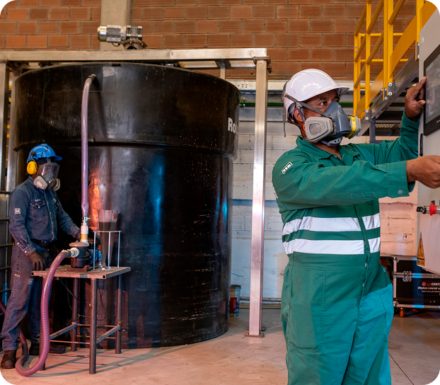 Dos operarios de Ciudad Limpia realizando labores de control y operación en el interior de una planta. En primer plano, un colaborador con uniforme verde, casco y respirador manipula un panel de control, mientras al fondo otro trabajador supervisa un tanque de gran tamaño conectado a mangueras y equipos del proceso.