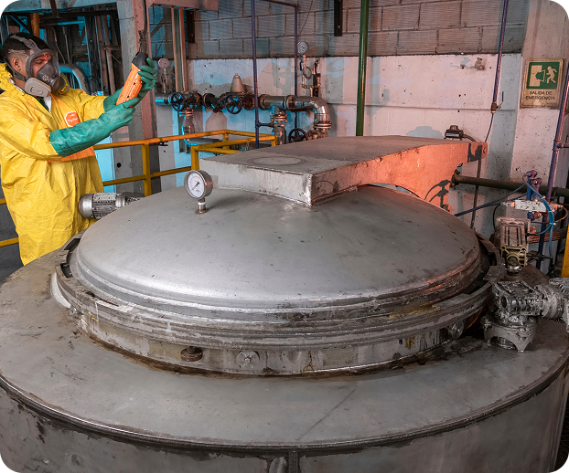 Colaborador de Ciudad Limpia, con traje de protección amarillo, guantes y respirador, inspeccionando o registrando información frente a un reactor o tanque metálico de gran tamaño en el interior de una planta. Alrededor se observan tuberías, válvulas, manómetros y barandas de seguridad propias del proceso de tratamiento.