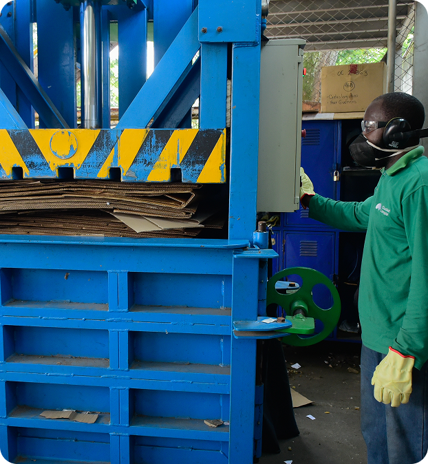 Colaborador de Ciudad Limpia opera una compactadora industrial de cartón en una planta. Usa guantes, gafas, protección auditiva y mascarilla, mientras supervisa el proceso de prensado de material dentro de una máquina azul.
