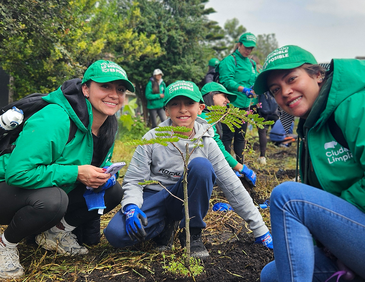 Grupo de voluntarios y colaboradores de Ciudad Limpia participando en una jornada de siembra al aire libre. En primer plano, dos mujeres y un niño posan sonrientes junto a un árbol recién plantado, mientras al fondo otras personas continúan las labores de restauración ambiental con guantes y gorras verdes institucionales.