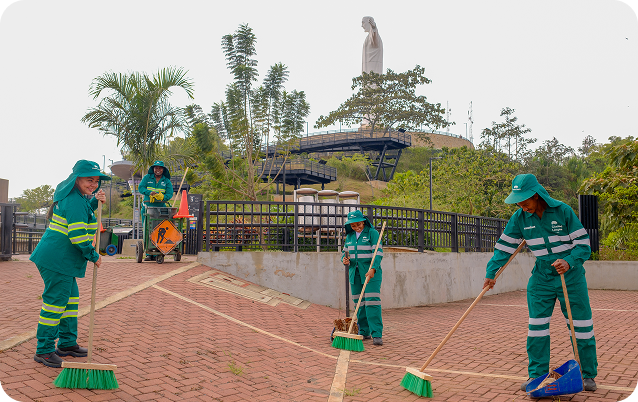 Cuatro colaboradoras de Ciudad Limpia realizan labores de barrido y recolección en la plazoleta de Cristo Rey, en Cali. Visten uniforme verde reflectivo y usan escobas y recogedor, mientras al fondo se observan jardines, barandas, un carro de aseo y un mirador con la estatua monumental en la parte alta del paisaje.